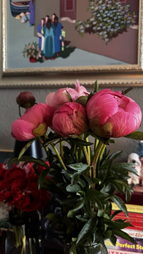Pink peonies in a vase on a shelf with books and a framed painting